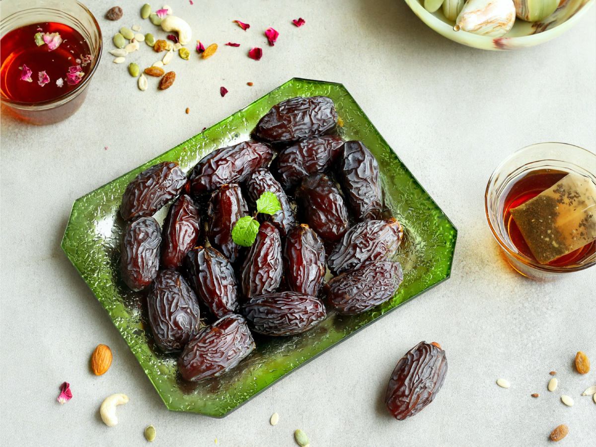 Top view of glossy Medjoul dates arranged on a green glass tray, garnished with mint leaves, and surrounded by herbal tea, dry fruits, and rose petals