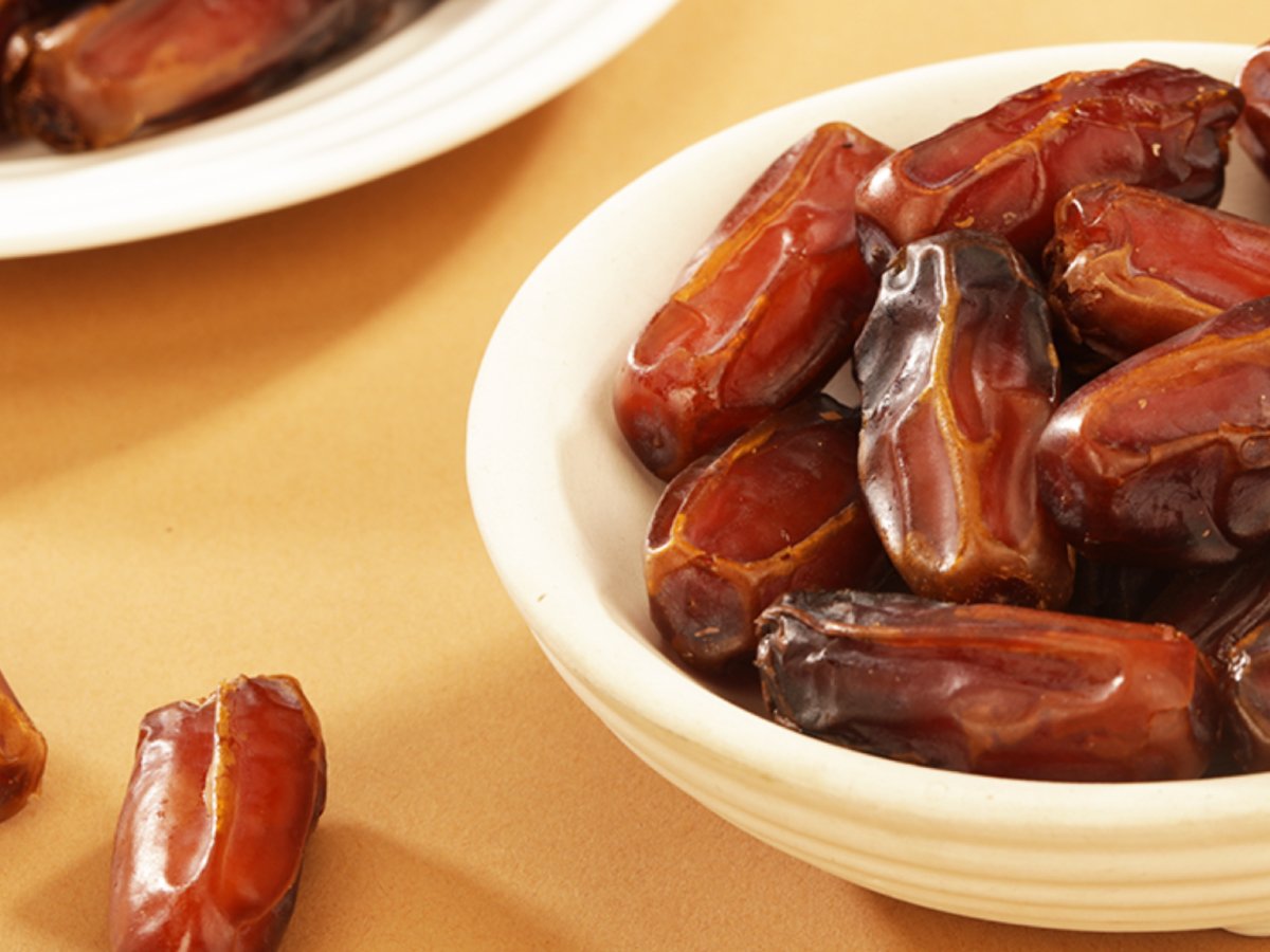 Close-up of Mabroom dates in a white ceramic bowl on a warm-toned surface, highlighting their firm texture, fibrous skin, and deep reddish-brown hue.